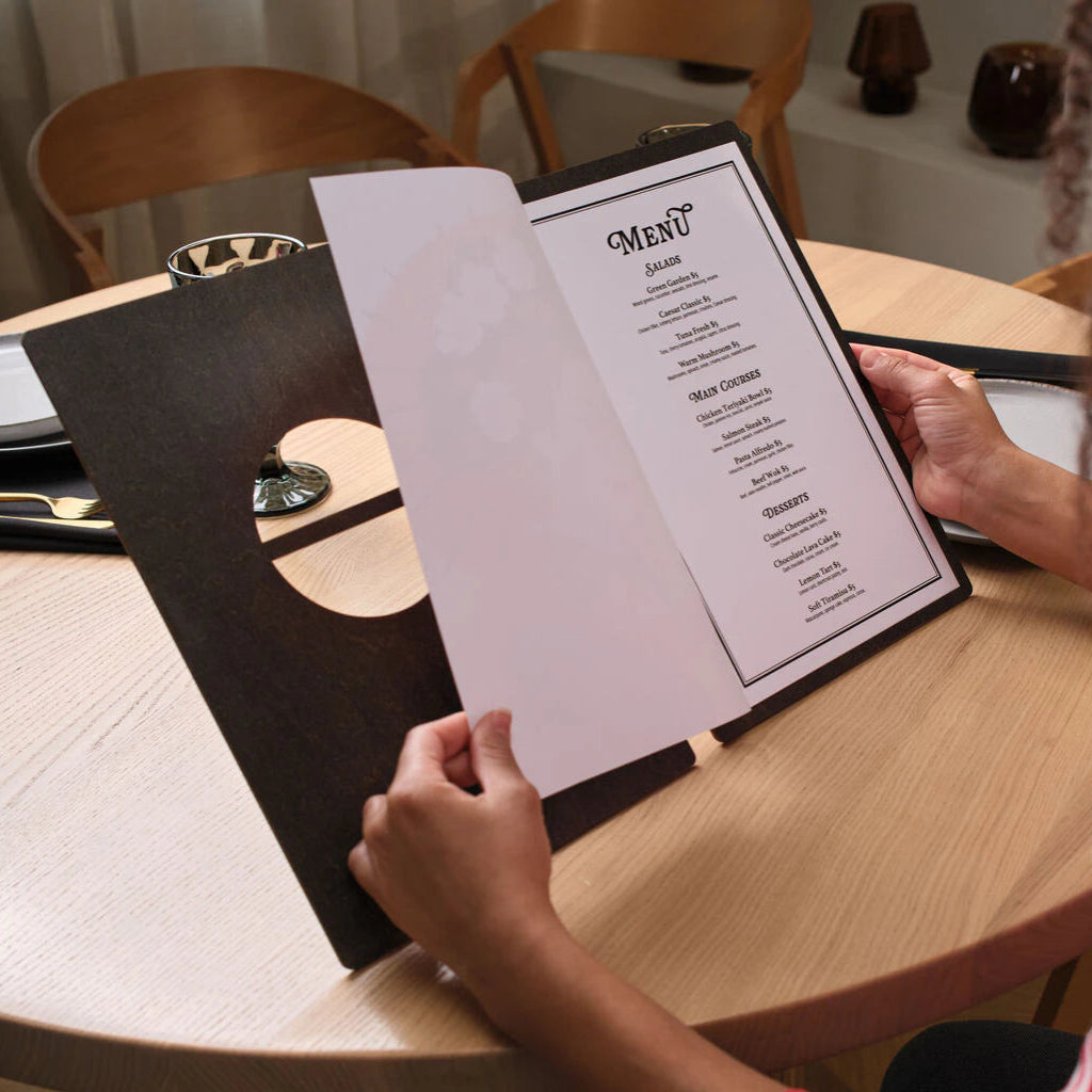 A top-down lifestyle shot of the fully opened wooden menu on a restaurant table, held by a hand to showcase the internal artwork alongside table settings like a glass and grapes.