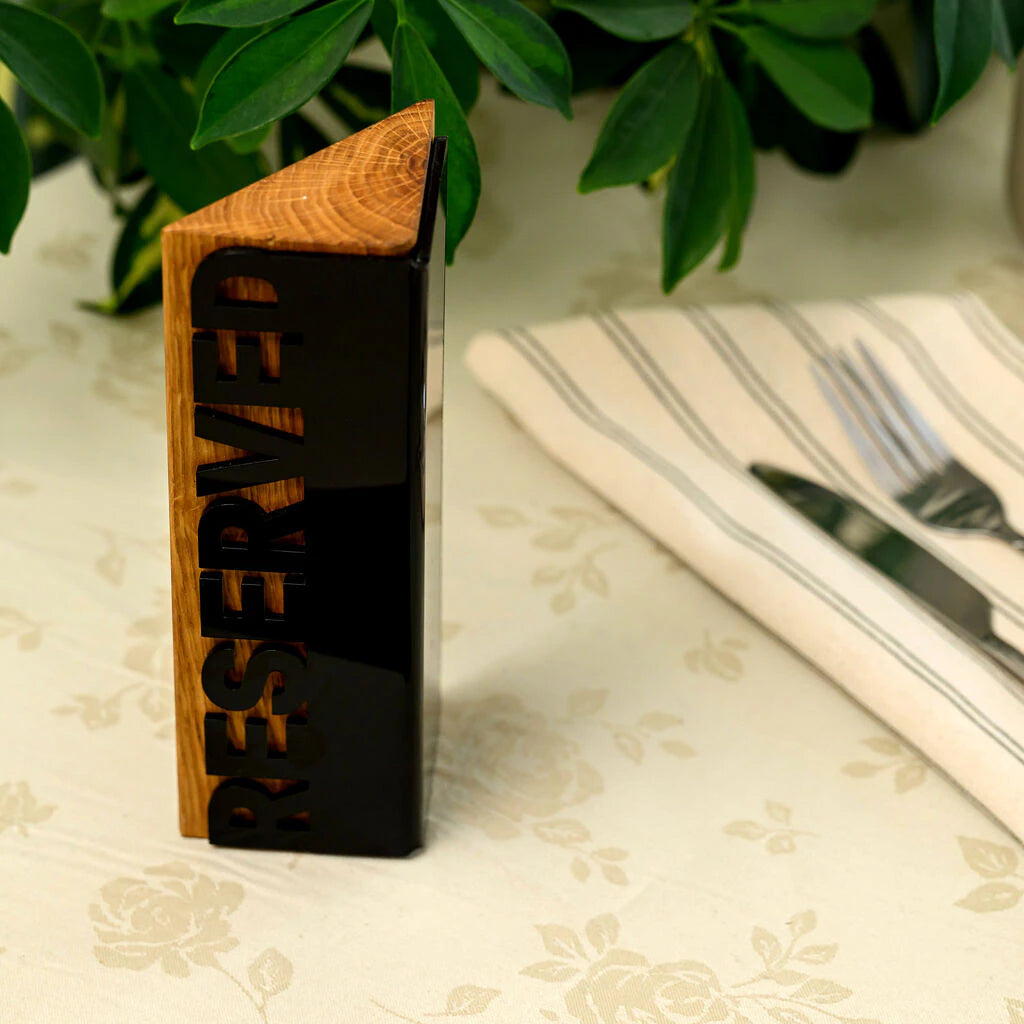 A modern 'Reserved' sign standing vertically on a floral tablecloth. The sign has a triangular prism shape made of natural wood, with a sleek black acrylic front panel featuring the word 'RESERVED' in cutout letters. A striped napkin with a knife and fork is visible in the blurred background