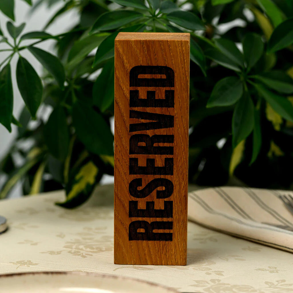 Front view of a vertical "RESERVED" table sign handcrafted from solid oak wood. The word is deeply laser-engraved in a bold, uppercase font, showcasing the beautiful natural grain and texture of the wood. The sign is placed on a set table in a restaurant with lush green plants in the background