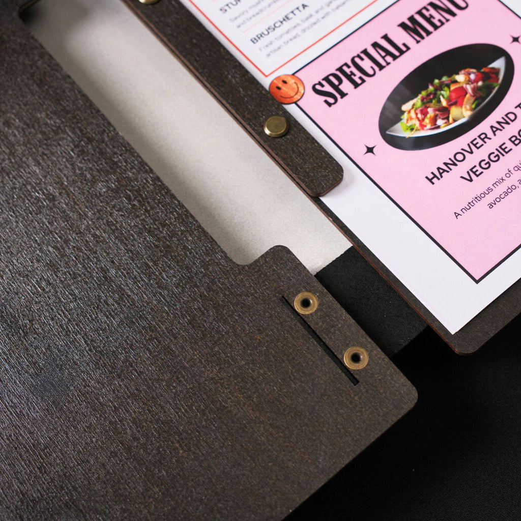 An interior close-up of an open wooden menu folder, showing a pink "Special Menu" page featuring a salad photo. The page is held in place by a dark wooden strip and brass binding hardware.