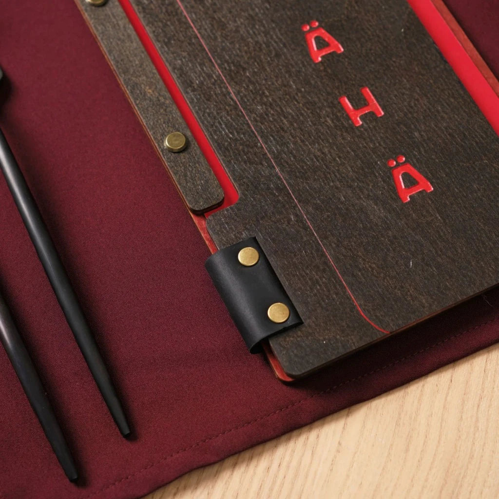 Close-up of a wooden menu folder with custom red color engraving and a wine bottle shaped cutout. Shows the premium dark wood finish, metal bolt binding, and a leather accent on a burgundy table setting