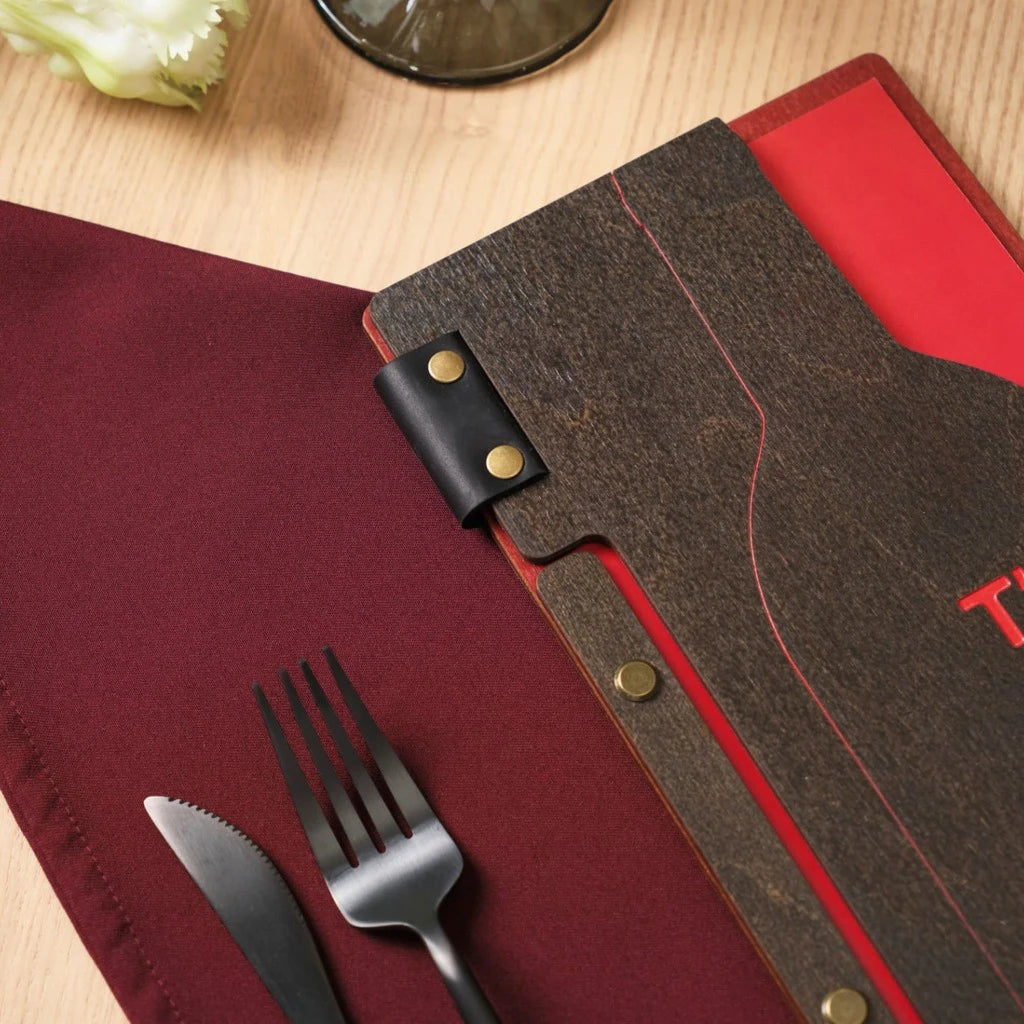 Close-up of a custom wood menu cover featuring a black leather hinge with gold bolts. This unique wooden menu holder showcases a wine bottle cutout and red color engraving on a burgundy napkin