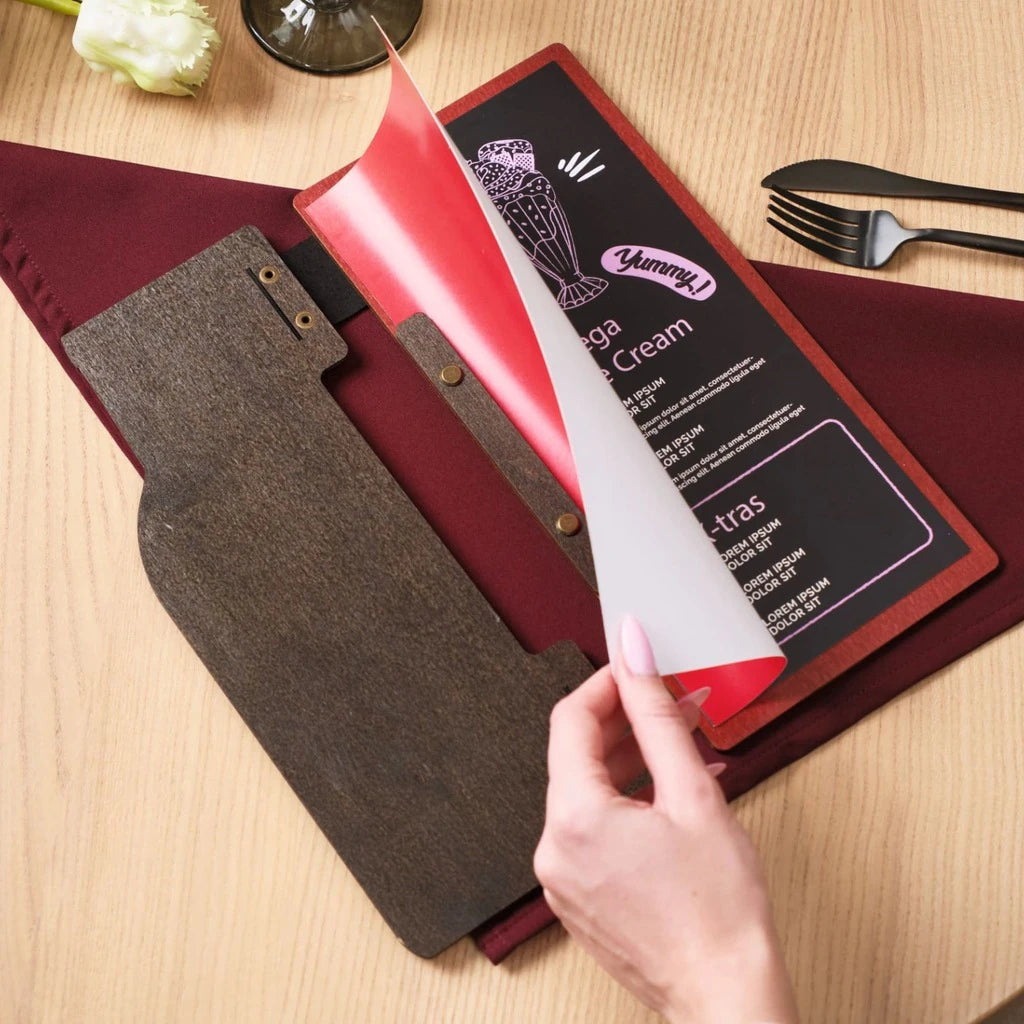 Demonstrating the easy page replacement in a custom wooden menu folder. Features a dark wood cover with a wine bottle cutout, bronze bolt binding, and a hand flipping a menu page over a red interior.