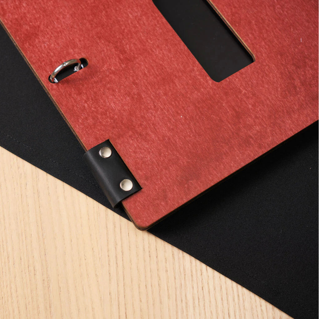 Macro shot of the bottom corner of a chili-red wooden menu, highlighting the black leather binding secured with silver metal rivets on a black fabric surface.