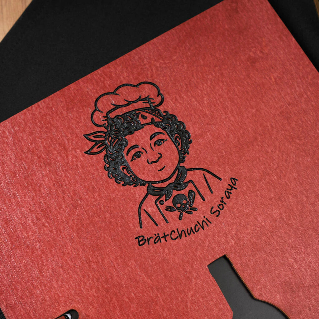 Detailed view of the black engraved logo featuring a stylized chef character and "Brätchuchi Soraya" text on the textured chili-red wooden menu surface.