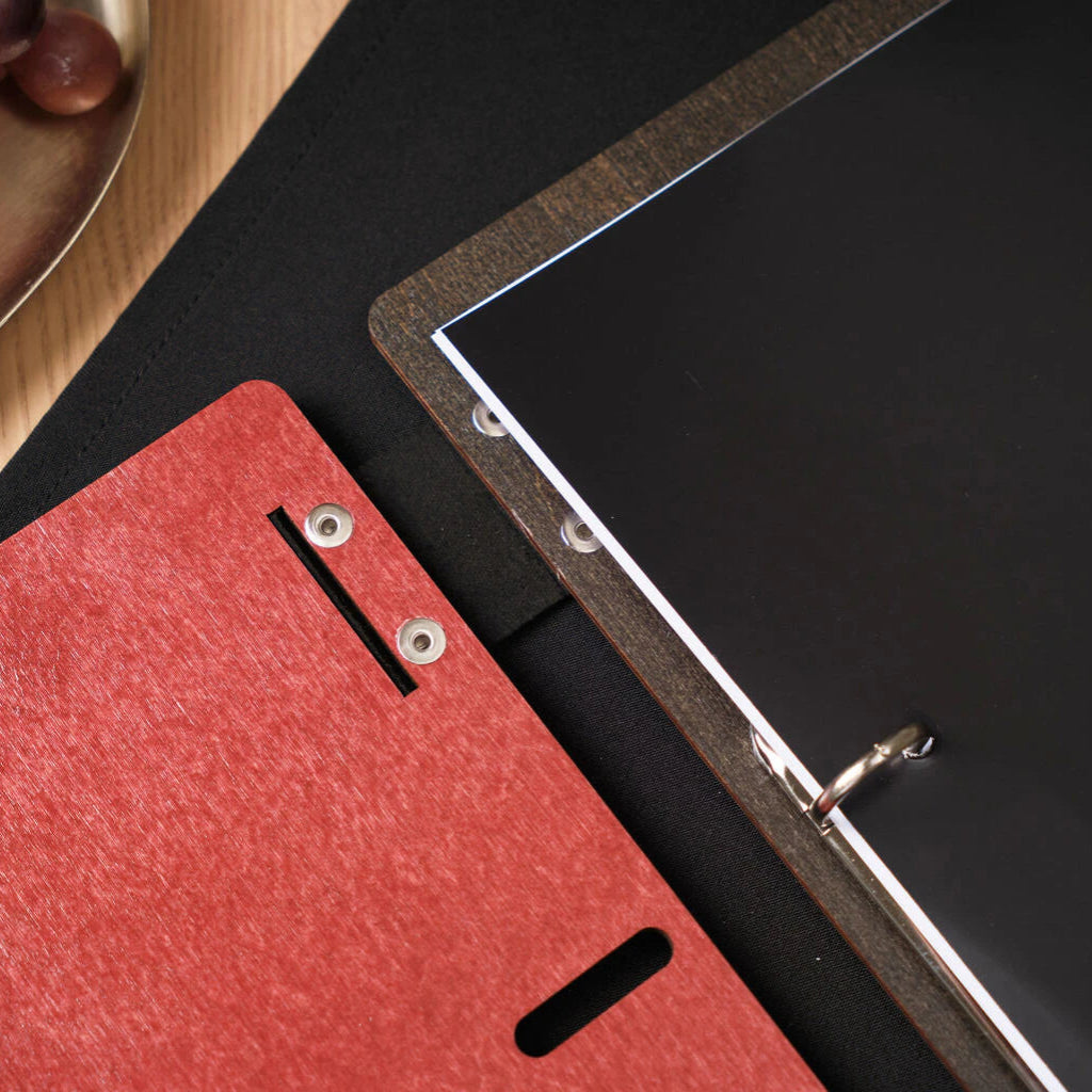 Close-up view of the spine of an open chili-red wooden menu, showing the metal ring binder and the contrast between the red exterior and dark interior.