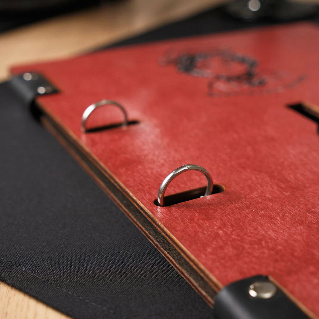 Close-up angle of a chili-red wooden menu folder, showing the silver ring mechanism and the layered edge of the wood on a black fabric surface